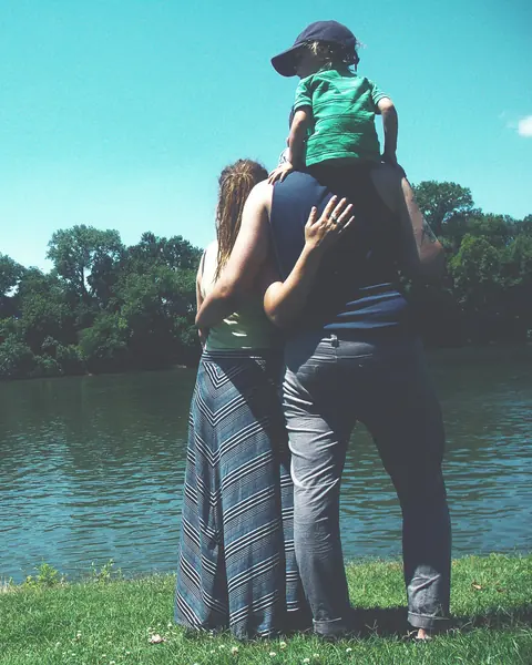 Un papà con il figlio sulle spalle e la mamma, di spalle, guardano un lago tra gli alberi