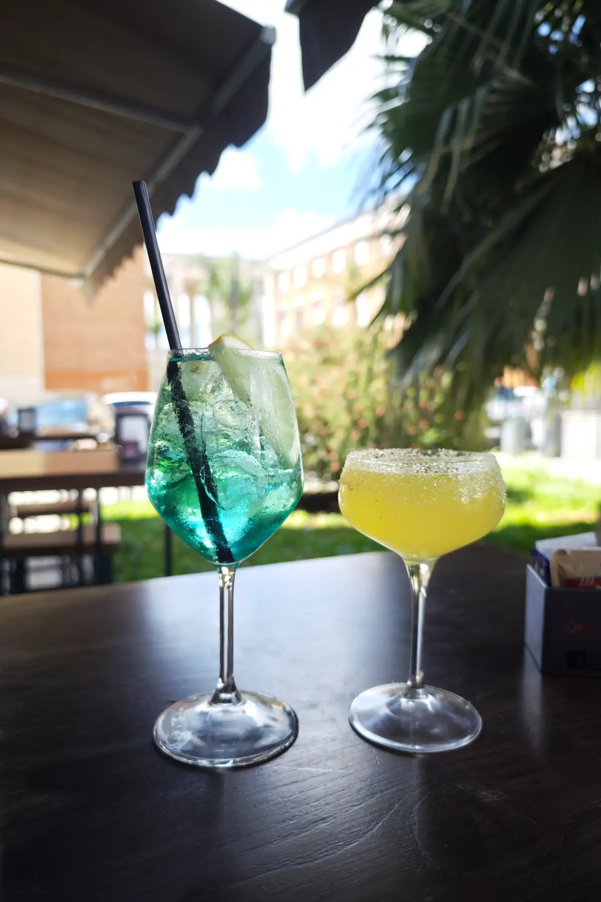 Uno spritz azzurro e un cocktail giallo sul tavolo all’aperto, fra le palme e la tenda del locale