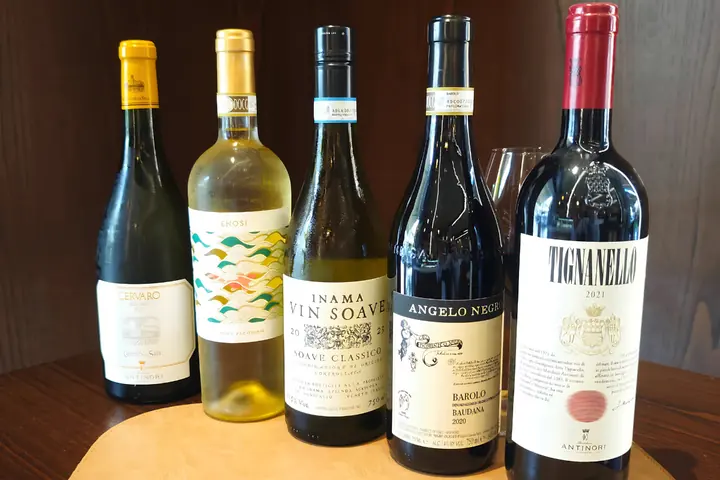 Cinque bottiglie di vino italiano in fila, tre bianchi e due rossi, con un calice accanto