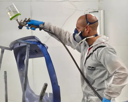 Un verniciatore in tuta e maschera spruzza il blu su un paraurti, nel forno della carrozzeria