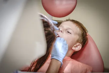 Un bambino sulla poltrona, a bocca aperta, mentre la dentista lo visita con lo specchietto