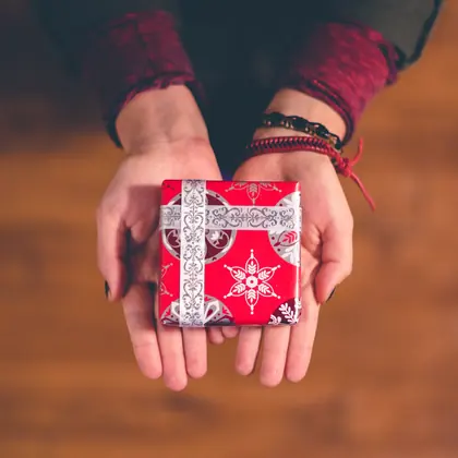 Due mani aperte porgono un piccolo regalo incartato di rosso, con il nastro argentato