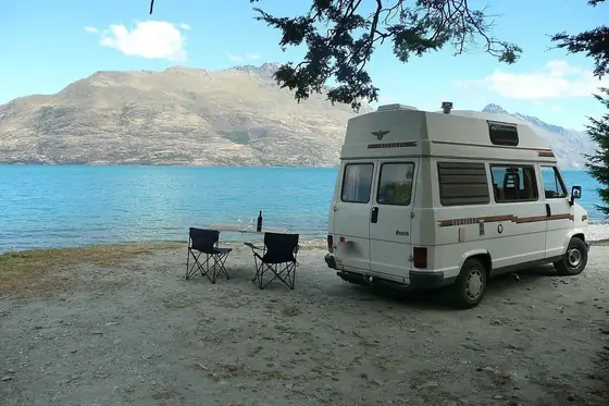 Camper parcheggiato in riva a un lago di montagna, con due sedie da campeggio