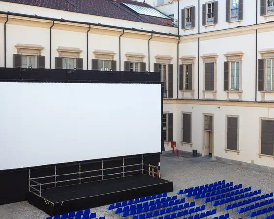 Un grande schermo per esterni montato nel cortile di un palazzo storico, davanti alle file di sedie