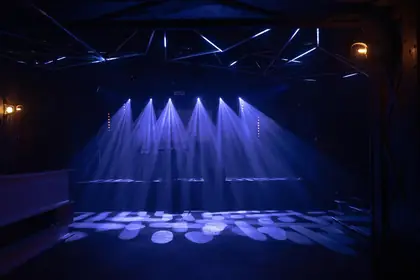 Sei fasci di luce blu che scendono su un palco vuoto, sotto un traliccio illuminato