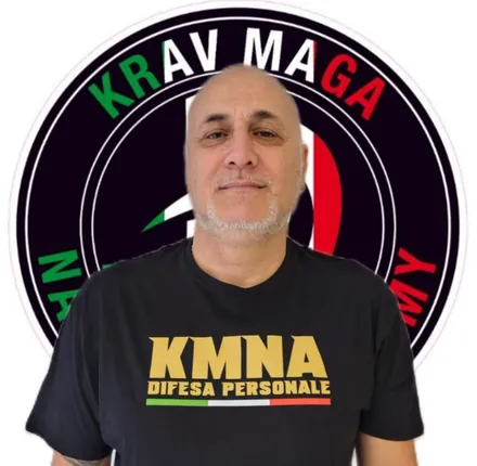 Un uomo sorridente con una maglietta nera da difesa personale, davanti a uno stemma del Krav Maga