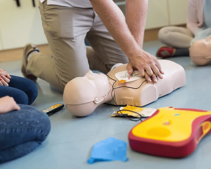 Un corso in aula: un istruttore in ginocchio fa il massaggio cardiaco su un manichino, accanto al defibrillatore, mentre gli allievi guardano