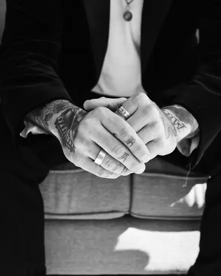 Le mani tatuate di un uomo in giacca scura, intrecciate sulle ginocchia