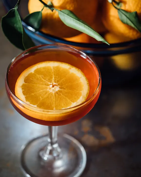 Un cocktail color ambra in una coppa con una fetta d'arancia, accanto a un cesto di arance