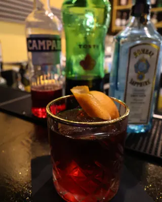 Un cocktail rosso con una scorza d’arancia, davanti alle bottiglie del bar
