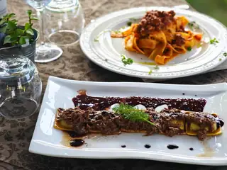 Pappardelle al ragù e un piatto di carne con una salsa scura, sulla tovaglia damascata