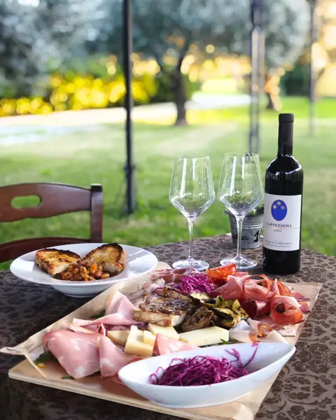 Una tavola in giardino fra gli ulivi, con un tagliere di salumi, due calici e una bottiglia di vino