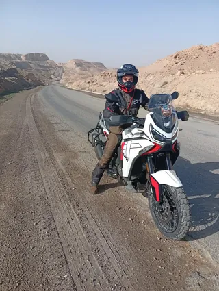 Un motociclista fermo con la sua moto da viaggio bianca, lungo una strada tra rocce e sabbia