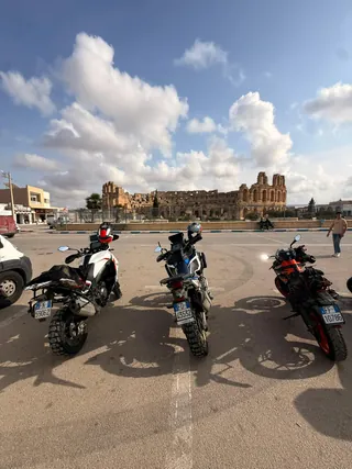 Tre moto parcheggiate in piazza davanti a un anfiteatro romano, sotto un cielo di nuvole