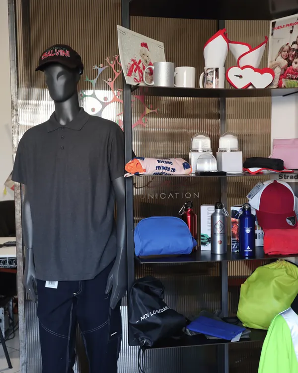 Un manichino con cappellino e polo, accanto agli scaffali di tazze, borracce, zaini e gadget personalizzati