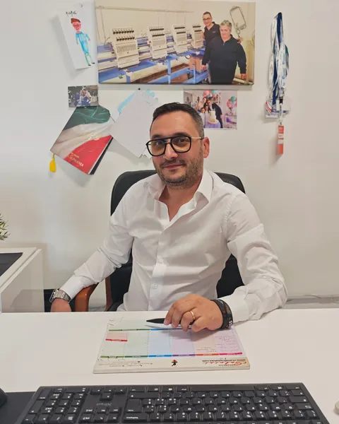 In camicia bianca alla scrivania dell’ufficio, con la foto delle ricamatrici appesa alle spalle