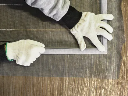 Due mani con i guanti da lavoro che tendono la rete di una zanzariera sul telaio