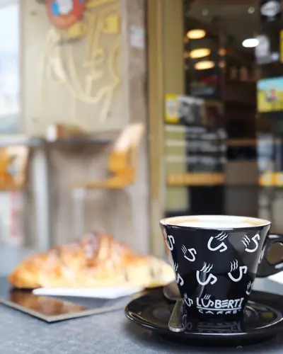 Cornetto e cappuccino in una tazza nera, sul tavolino all’ingresso del bar